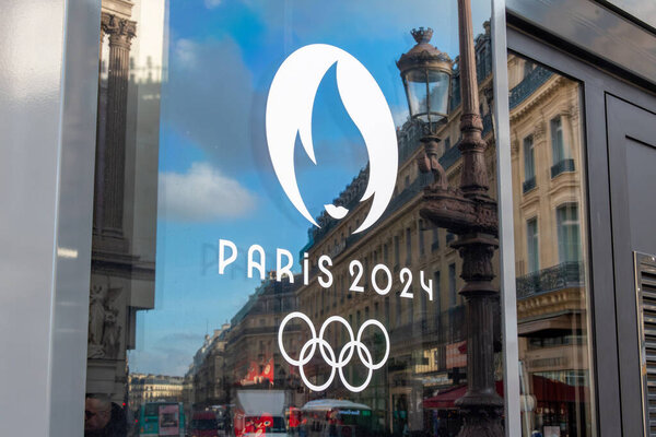 Paris, France - December 6, 2023: Close-up of the Paris 2024 logo, symbol of the Summer Olympic Games of 2024 in Paris, on a window with the reflection of a Paris street