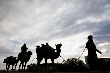 Khiva, Özbekistan - 11 Temmuz 2024: Güneş batarken bir deve kervanı ve rehberini tasvir eden bronz heykellerin siluetleri İpek Yolu yolculukları ve göçebe gelenekleri için çarpıcı bir övgü niteliğindedir..
