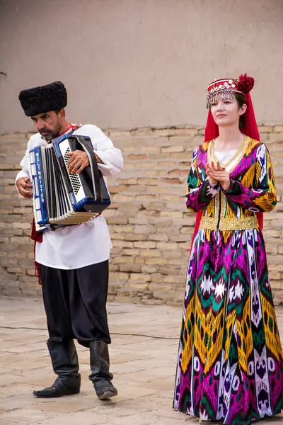 Khiva, Özbekistan - 11 Temmuz 2024: Khiva 'nın tarihi merkezinde geleneksel Özbek halk performansı. Renkli ulusal elbiseli bir kadın mikrofona şarkı söylerken, diğeri arkasında duruyor ve bir erkek akordeon çalıyor. Hepsi çok şık giyinmiş.