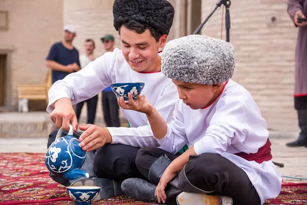 Khiva, Özbekistan - 11 Temmuz 2024: Bir kültür etkinliği sırasında geleneksel giyinmiş Özbek çocuklar. Biri çay koyarken diğeri Orta Asya 'daki konukseverliğin temsilini izliyor..