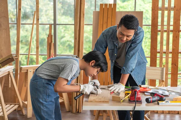 Asian boy is driving nails into plank with his carpenter father nearby. It\'s family activity on vacation concept.