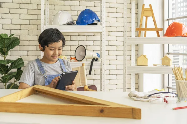 Asian boy is studying woodworking on tablet with various tools scattered on wooden table.