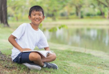 Şirin Asyalı çocuk mutlu, bahçede çimlerin üzerinde oturuyor ve okuyor. Arka planda bulanık yapraklar var..
