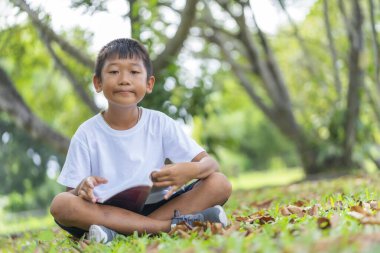 Şirin Asyalı çocuk mutlu, bahçede çimlerin üzerinde oturuyor ve okuyor. Arka planda bulanık yapraklar var..