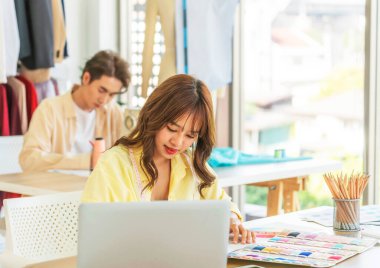 Çekici kadın terzi ve tasarımcı masada mutlu bir şekilde oturuyor. Çekici erkek arkadaşı arkasında oturuyor ve giysileri çamaşır ipine asılı. Masadaki kağıt ve defter bilgisayarları.