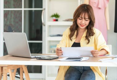 Çekici kadın terzi ve tasarımcı. Masasında mutlu bir şekilde oturuyor ve gülümsüyor. Mankenler arkasında asılı duruyor. Masanın üzerinde bir kağıt ve defter var..