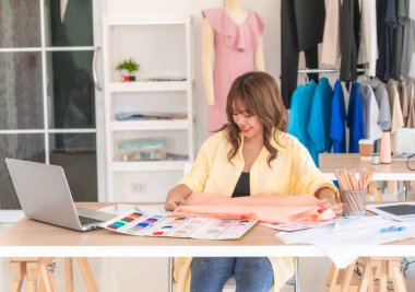 Çekici kadın terzi, bantla ölçü alarak masanda dikiş aletleri, renk şemaları ve dizüstü bilgisayarla mutlu bir şekilde kumaşı ölçüyor..