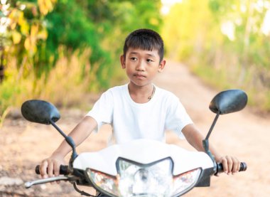 Şirin ve yaramaz çocuk bahçede toprak yolda motosiklet sürmeyi öğreniyor. Arka planda bulanık yeşil ve sarı yapraklar var..