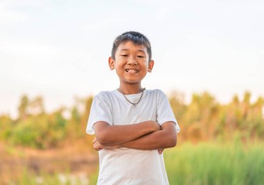 Parkta kollarını kavuşturmuş haylaz bir çocuğun portresi. Arka planda bulanık ağaç çizgileriyle toprak yolda mutlu bir şekilde gülümsüyor. Sonbahar konsepti.