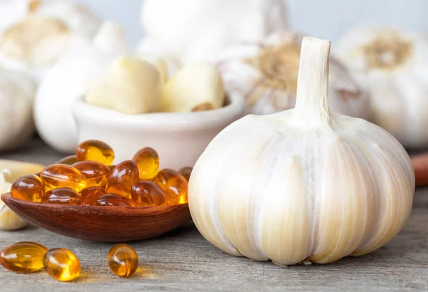Garlic bulbs, capsules oil in wooden spoon, garlic cloves in white bowl, and on old wooden table with blurred white wooden background. Garlic reduce risk of many diseases. Healthy food concept.