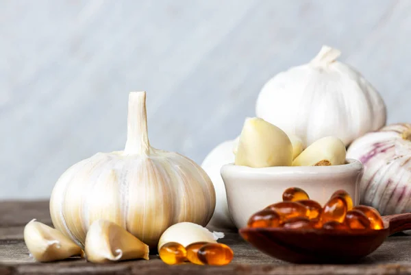 Garlic bulbs, capsules oil in wooden spoon, garlic cloves in white bowl, and on old wooden table with blurred white wooden background. Garlic can help reduce risk of many diseases. Healthy food concept.