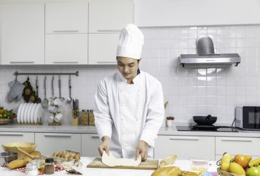 Şapkalı ve önlüklü hamurlu bir erkek şef portresi. Evde mutfakta hazırlanırken. Beyaz önlüklü Asyalı şef mutfaktaki masada hamur işi yapıyor.. 