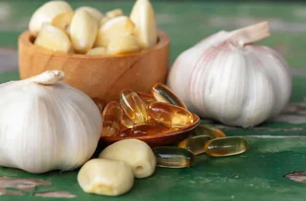 Gold garlic extract oil capsules in wooden spoon, garlic bulbs, and garlic cloves in brown bowl, and on old green wooden background. Garlic can help reduce the risk of many diseases. Classified as healthy food.