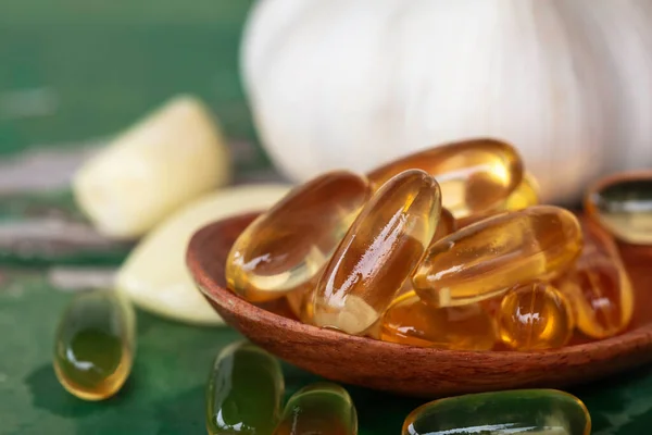 Close up of gold garlic extract capsules oil in wooden spoon, garlic bulbs, and garlic cloves in brown bowl, and on old green wooden background. Garlic can help reduce the risk of many diseases.
