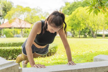 Sağlıklı bir kadın, güneşli bir sabahta bulanık ağaç arkaplanlı bir parkta vücudunu güçlendirmek için spor kıyafetleri giyer..