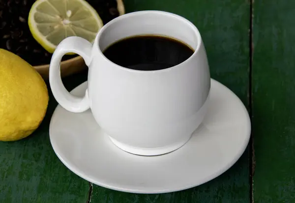 Hot black coffee in white cup, with yellow lemon, coffee beans in wooden saucer and lemon slices on old green wooden background.
