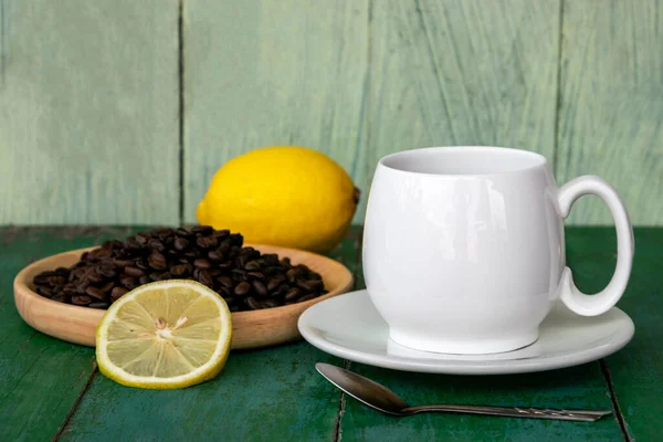 Hot black coffee in white cup, with yellow lemon, coffee beans in wooden saucer and lemon slices on old green wooden background.