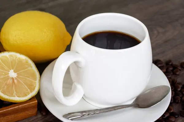 Warm black coffee in white cup with yellow lemon, coffee beans, and lemon slices on vintage brown wooden background. Healthy drink concept.