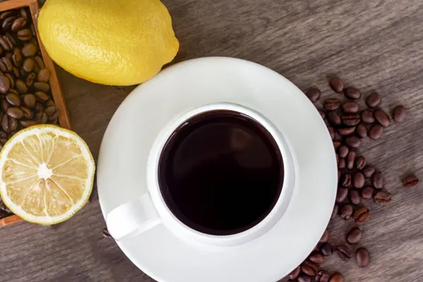 Top view of warm black coffee in white cup with yellow lemon, coffee beans, and lemon slices on vintage brown wooden background. Healthy drink concept.