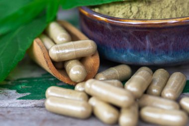 Paniklemenin ahşap kaşık içindeki bitkisel kapsülleri (Andrographis Paniculata) ve seramik kasede toz özü bulunan yeşil yapraklar eski ahşap arka planda..