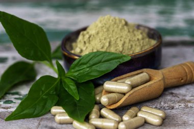 Paniklemenin ahşap kaşık içindeki bitkisel kapsülleri (Andrographis Paniculata) ve seramik kasede toz özü bulunan yeşil yapraklar eski ahşap arka planda..