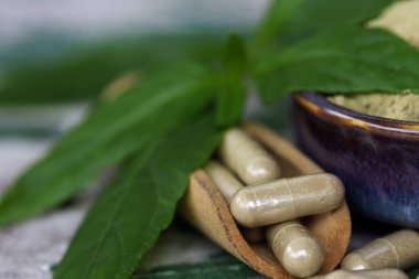 Paniklemenin ahşap kaşık içindeki bitkisel kapsülleri (Andrographis Paniculata) ve seramik kasede toz özü bulunan yeşil yapraklar eski ahşap arka planda..