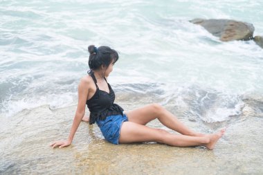 Woman Sitting on Sea Stone Beach at Coast,Happy Beauty Girl Enjoy with Blue Ocean,Authentic Skin Tan,Black Hair,Slim fit Body Asian Thailand,Tourism Relax Vacation Tropical Travel Holidays Nature.