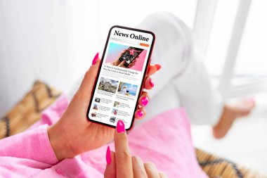 Evde oturan ve cep telefonundan haber okuyan bir kadın.