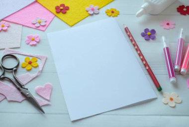 Renkli keçeli çiçekler, makas, yapıştırıcı ve boş kağıt da dahil olmak üzere DIY tebrik kartı el işi malzemelerinin yaratıcı düz konumu, beyaz ahşap bir arka planda düzenlenmiş, özel bir el yapımı kart yapma sürecini gösteren 