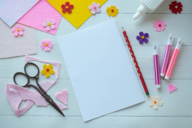 Renkli keçeli çiçekler, makas, yapıştırıcı ve boş kağıt da dahil olmak üzere DIY tebrik kartı el işi malzemelerinin yaratıcı düz konumu, beyaz ahşap bir arka planda düzenlenmiş, özel bir el yapımı kart yapma sürecini gösteren 