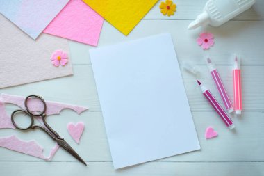 Renkli keçeli çiçekler, makas, yapıştırıcı ve boş kağıt da dahil olmak üzere DIY tebrik kartı el işi malzemelerinin yaratıcı düz konumu, beyaz ahşap bir arka planda düzenlenmiş, özel bir el yapımı kart yapma sürecini gösteren 
