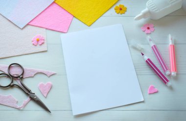 Renkli keçeli çiçekler, makas, yapıştırıcı ve boş kağıt da dahil olmak üzere DIY tebrik kartı el işi malzemelerinin yaratıcı düz konumu, beyaz ahşap bir arka planda düzenlenmiş, özel bir el yapımı kart yapma sürecini gösteren 