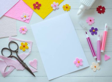 Renkli keçeli çiçekler, makas, yapıştırıcı ve boş kağıt da dahil olmak üzere DIY tebrik kartı el işi malzemelerinin yaratıcı düz konumu, beyaz ahşap bir arka planda düzenlenmiş, özel bir el yapımı kart yapma sürecini gösteren 