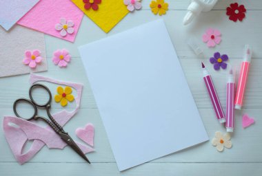Renkli keçeli çiçekler, makas, yapıştırıcı ve boş kağıt da dahil olmak üzere DIY tebrik kartı el işi malzemelerinin yaratıcı düz konumu, beyaz ahşap bir arka planda düzenlenmiş, özel bir el yapımı kart yapma sürecini gösteren 