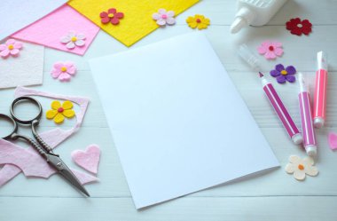 Renkli keçeli çiçekler, makas, yapıştırıcı ve boş kağıt da dahil olmak üzere DIY tebrik kartı el işi malzemelerinin yaratıcı düz konumu, beyaz ahşap bir arka planda düzenlenmiş, özel bir el yapımı kart yapma sürecini gösteren 