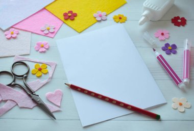 Renkli keçeli çiçekler, makas, yapıştırıcı ve boş kağıt da dahil olmak üzere DIY tebrik kartı el işi malzemelerinin yaratıcı düz konumu, beyaz ahşap bir arka planda düzenlenmiş, özel bir el yapımı kart yapma sürecini gösteren 