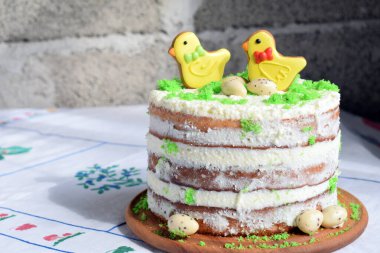 Sarı civciv şeklinde zencefilli kurabiyelerle kaplı çıplak katlı pasta, yeşil şeker parçacıklarıyla süslenmiş, ahşap arka planda geleneksel Paskalya kutlaması için bayram kırsal tatlı.