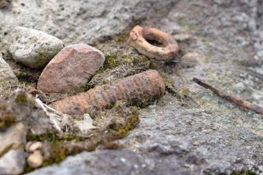 Paslı çivi ve paslı metal kalıntıları yosunlu zeminde kayaların arasında duruyor. Potansiyel yaralanma riskini ve terk edilmiş alandaki tetanos enfeksiyonunu vurguluyor.