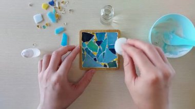 Rengarenk camlı mozaik bardak altlığı yüzeyindeki fazla beyaz alçıyı, el yapımı DIY işçiliği sırasında süngerle harç dolduktan sonra parçaları temizlemek için temizliyorum.