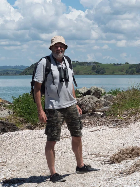 A smiling bearded nature loving man in camouflage shorts has binoculars and a ruck sack as he stands on a white shell beach in New Zealand
