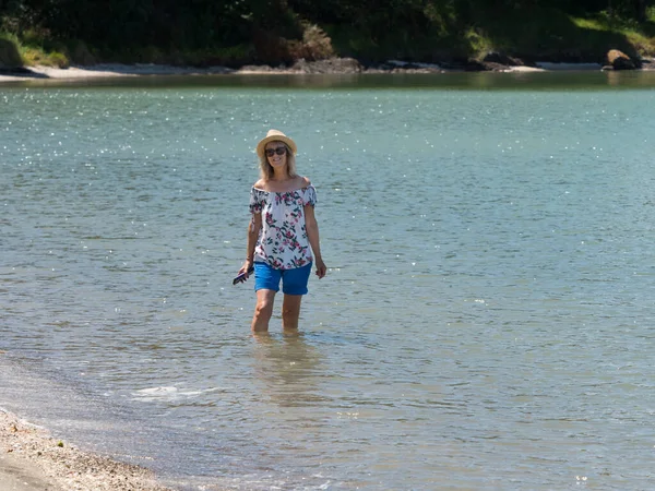 stock image An attractive lady smiles in shorts and sun hat as she walks and paddles in sea water in a New Zealand coastal bay