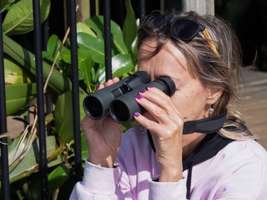 Bir kadın dürbünle bakar. Kafasında mor oje ve güneş gözlüğü vardır.