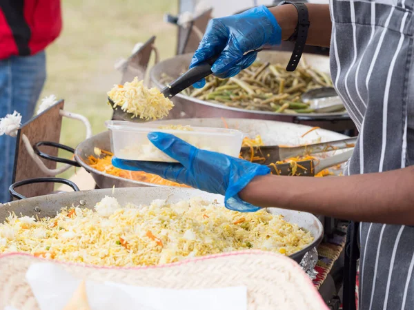 A store holder at a festival dishes up food into a plastic container wearing blue gloves to avoid cross infection. Arms and hands can be seen with pans of rice carrot and greens visible
