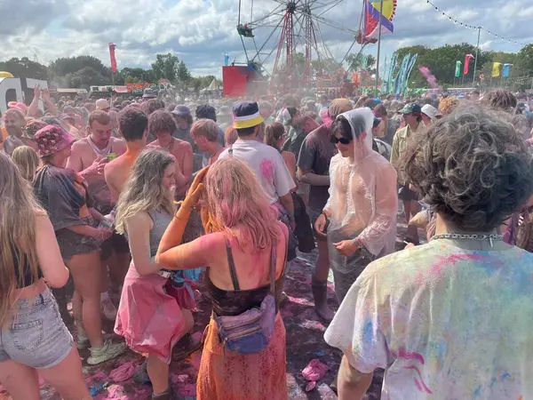 Stock image Oxford-UK-July 23,2023: A Festival crowd have been in a paint bomb event.Young people covered in multiple colours of powdered paint also on the ground.Big wheel, flags in background