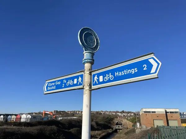 A sign indicates the cycle route and walkway section of a coastal path from Glyne Gap, Bexhill to Hastings.Mileages are shown. Blue sign against and sunny blue sky