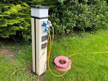 Birkaç kabloyu takılı bir elektrik bağlantı noktası ve yerde bir kablo makarası. Gece lambası yerleştirilmiş. Arkaplanda yeşil çit var.