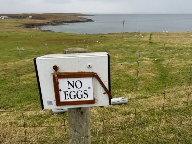 A box attached to a fence has a sign saying 'No Eggs'.Background a rugged coastal scene with sea . clipart