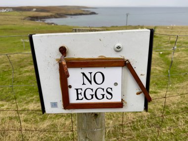 A box attached to a fence post has a sign saying 'No Eggs'.Background a rugged coastal scene with sea  clipart