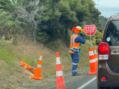 Yol kenarında çalışan bir işçi durup trafiği kontrol etmek için gemiye biniyor. Turuncu hi viz giysileri ve güneş şapkası giyiyorlar ve onları korumak için turuncu ve beyaz konileri var..