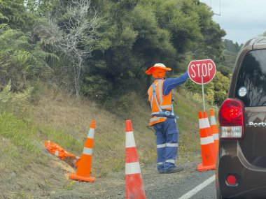 Yol kenarında çalışan bir işçi durup trafiği kontrol etmek için gemiye biniyor. Turuncu hi viz giysileri ve güneş şapkası giyiyorlar ve onları korumak için turuncu ve beyaz konileri var. .